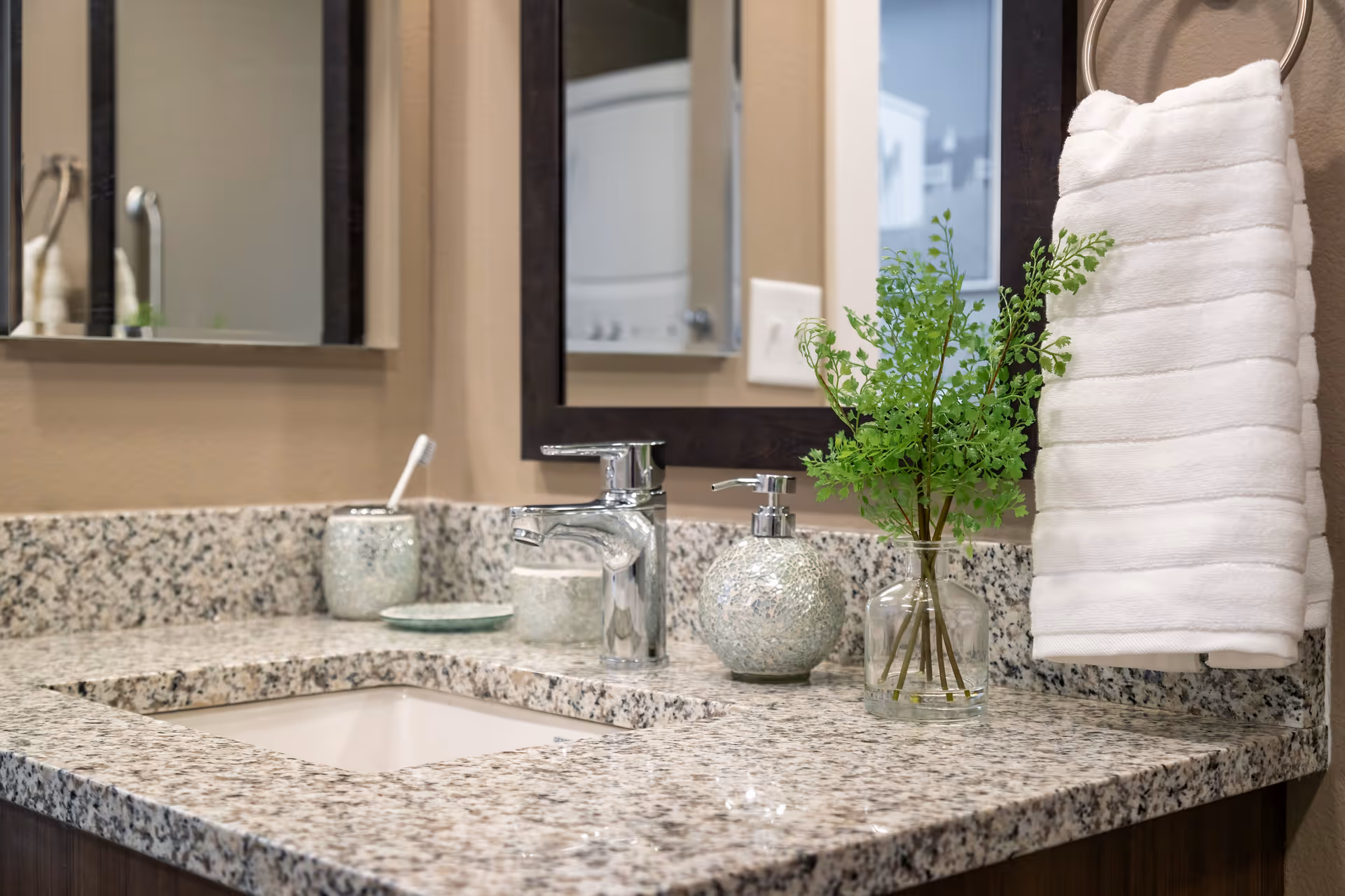 A bathroom countertop with a granite surface featuring a chrome faucet, a soap dispenser, a toothbrush holder with a toothbrush, a small dish, a glass vase with green leafy stems, and a white towel hanging on a ring next to a mirror.