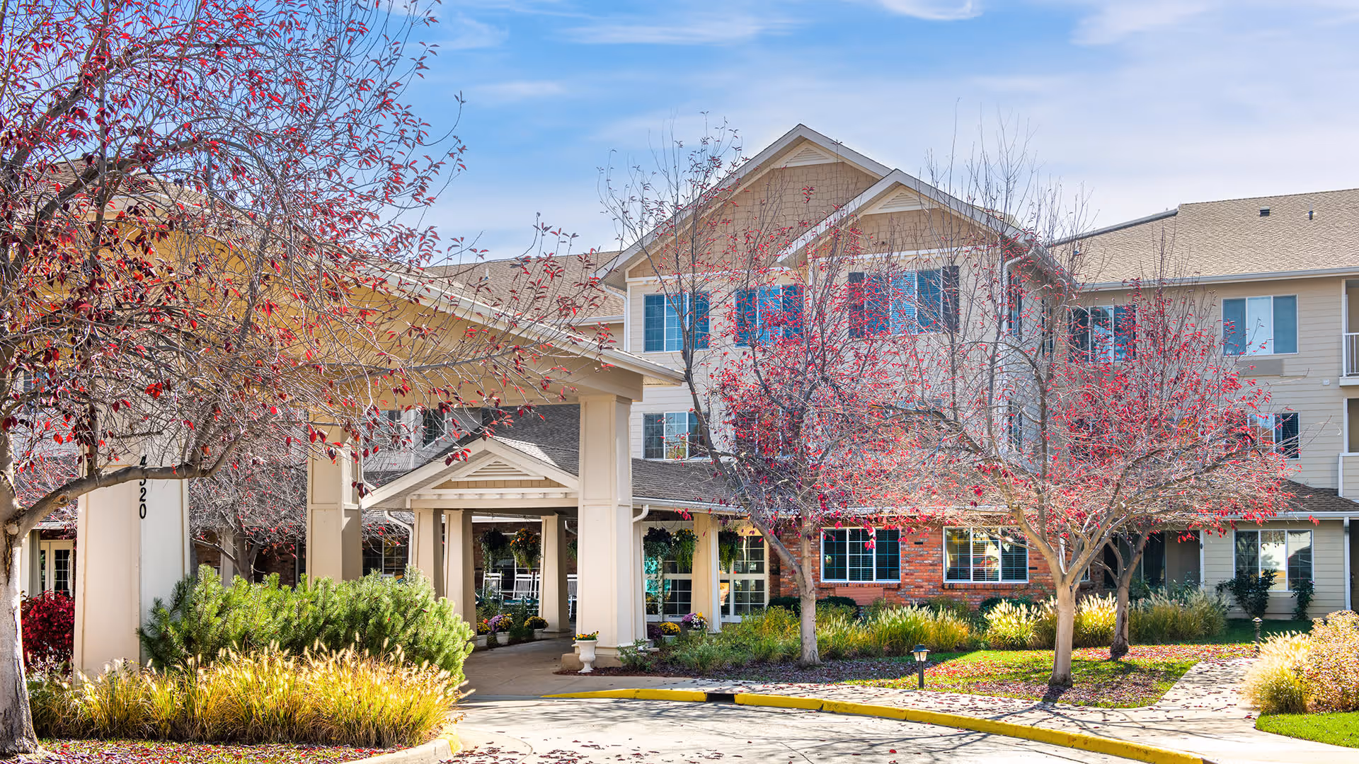 Exterior view of Sugar Valley Estates | Sky Active Living, showing a multi-story senior living facility with beige siding and brick accents. The entrance is covered by a large portico, surrounded by landscaped greenery and trees with red leaves under a blue sky.