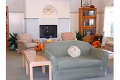 A cozy living room area with a green upholstered loveseat in the foreground, two patterned armchairs, a wooden side table with a floral arrangement, a fireplace with a decorative tile surround, and a wooden bookshelf filled with books and decor items.