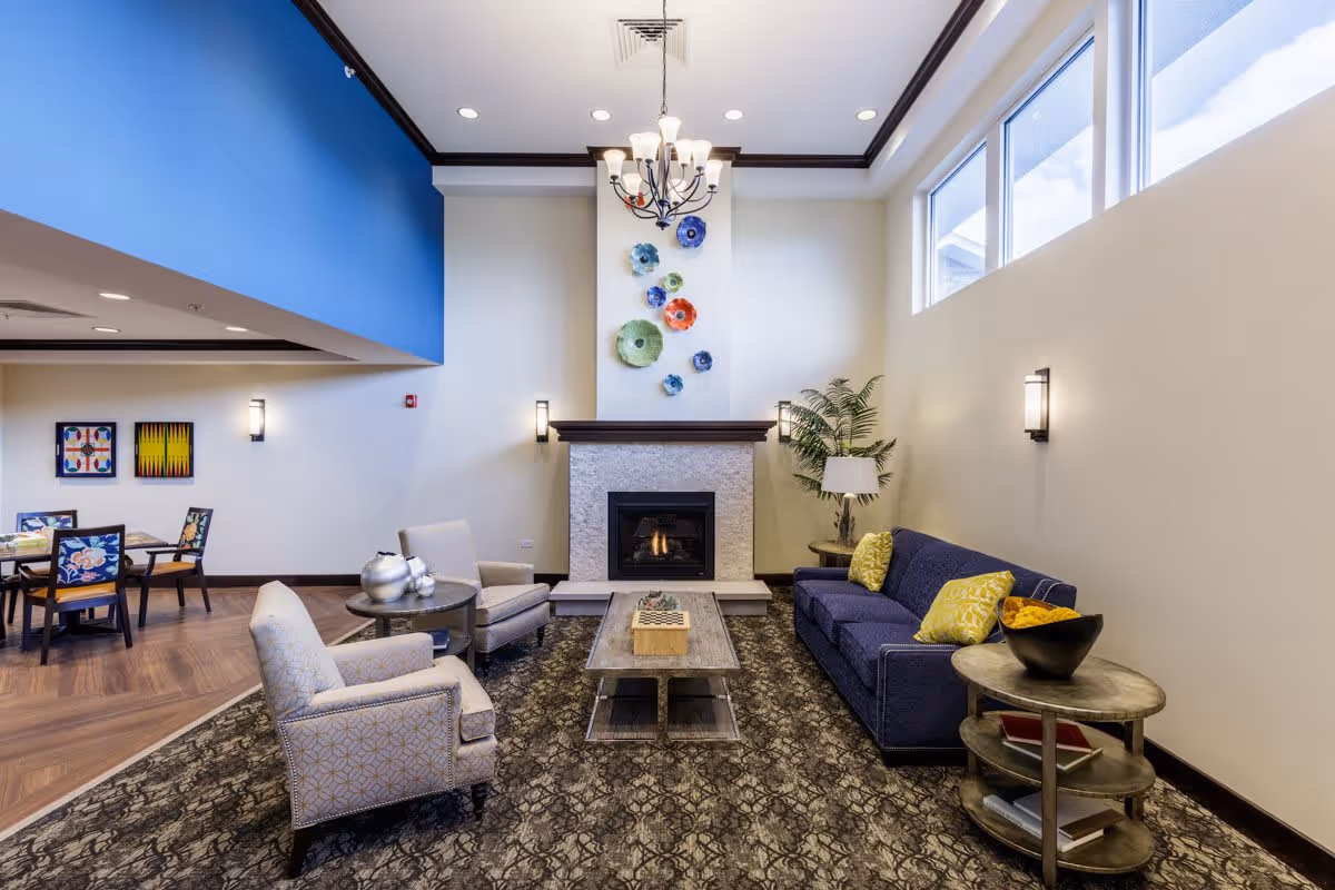 A cozy living room area with a blue sofa adorned with yellow pillows, two patterned armchairs, a wooden coffee table, and a side table with books and a decorative bowl. The room features a fireplace with colorful decorative plates mounted above it, a chandelier hanging from the ceiling, and large windows letting in natural light. In the background, there is a dining area with tables and chairs, and framed artwork on the wall.