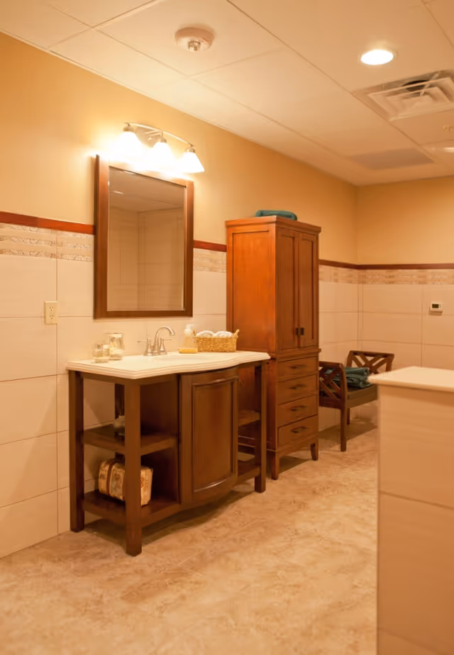 A bathroom interior featuring a wooden vanity with a white countertop and a sink, a rectangular mirror with a wooden frame above the sink, a tall wooden cabinet with drawers and doors, and a wooden chair with folded towels on it. The walls are tiled halfway up with a decorative border, and the floor has a light brown tile. The lighting is warm and soft.