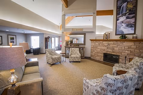 A spacious living room area with beige carpet, a stone fireplace, and several upholstered chairs and sofas arranged for conversation. The room features high ceilings with wooden beams, large windows allowing natural light, and framed artwork on the walls.