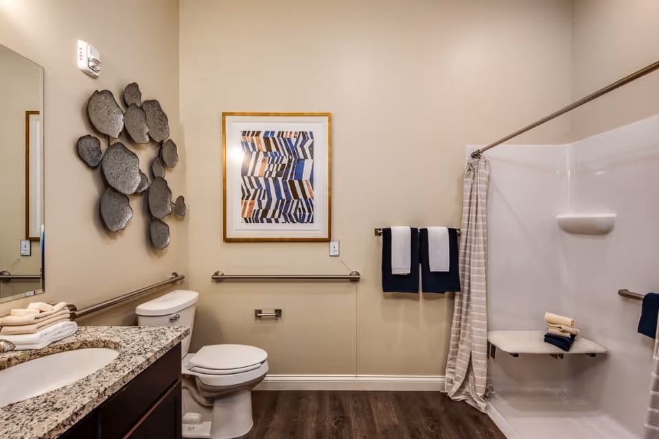 A clean and accessible bathroom with beige walls and dark wood flooring. The room features a granite countertop with a sink, a toilet with grab bars on both sides, a walk-in shower with a fold-down seat and a striped shower curtain. Two navy blue and white towels hang on a towel rack next to the shower. There is a framed abstract artwork on the wall above the toilet and decorative metal wall art on the adjacent wall.