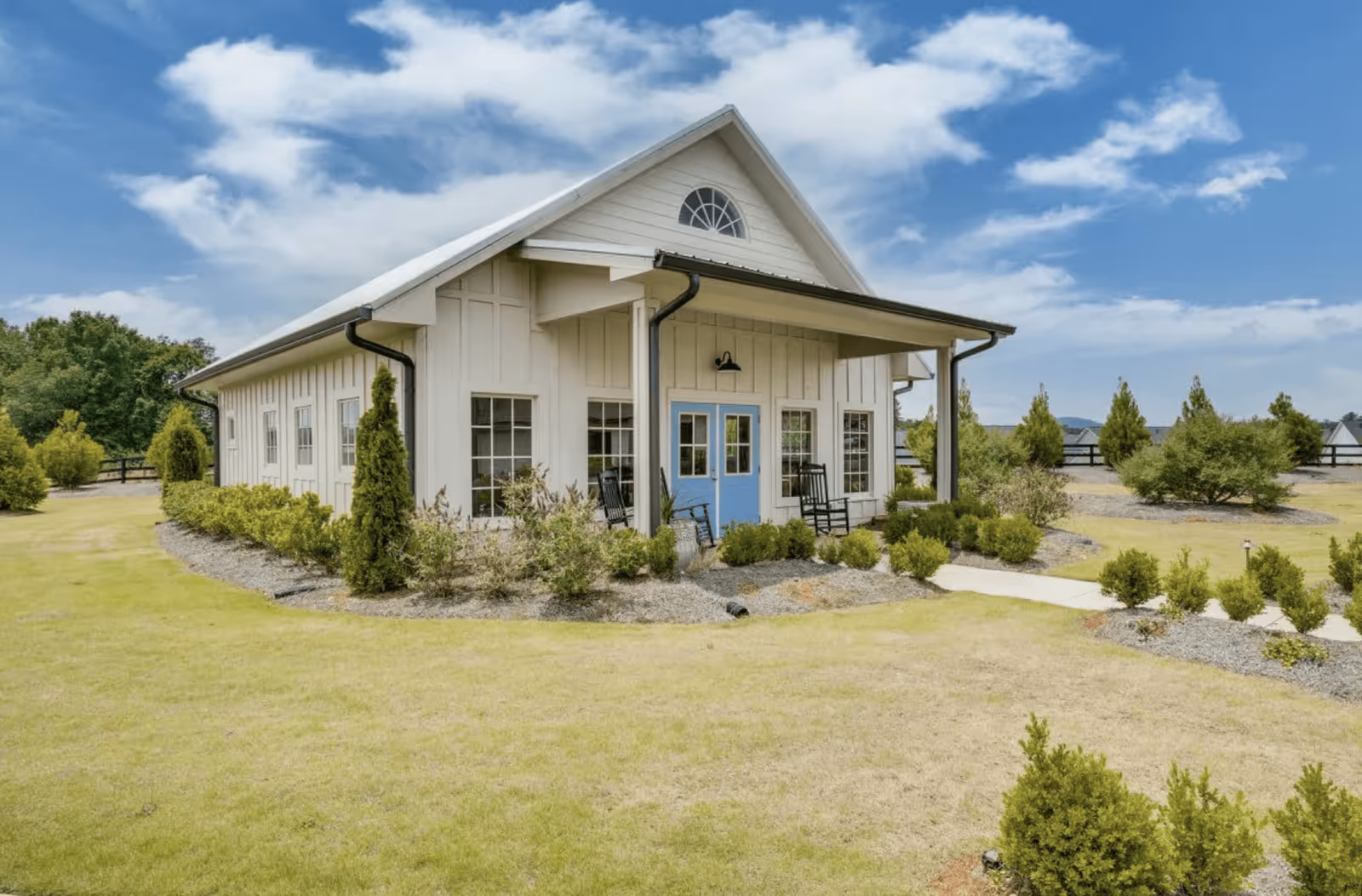 A single-story building with white siding and a blue double door entrance, surrounded by neatly trimmed bushes and a well-maintained lawn under a partly cloudy sky.