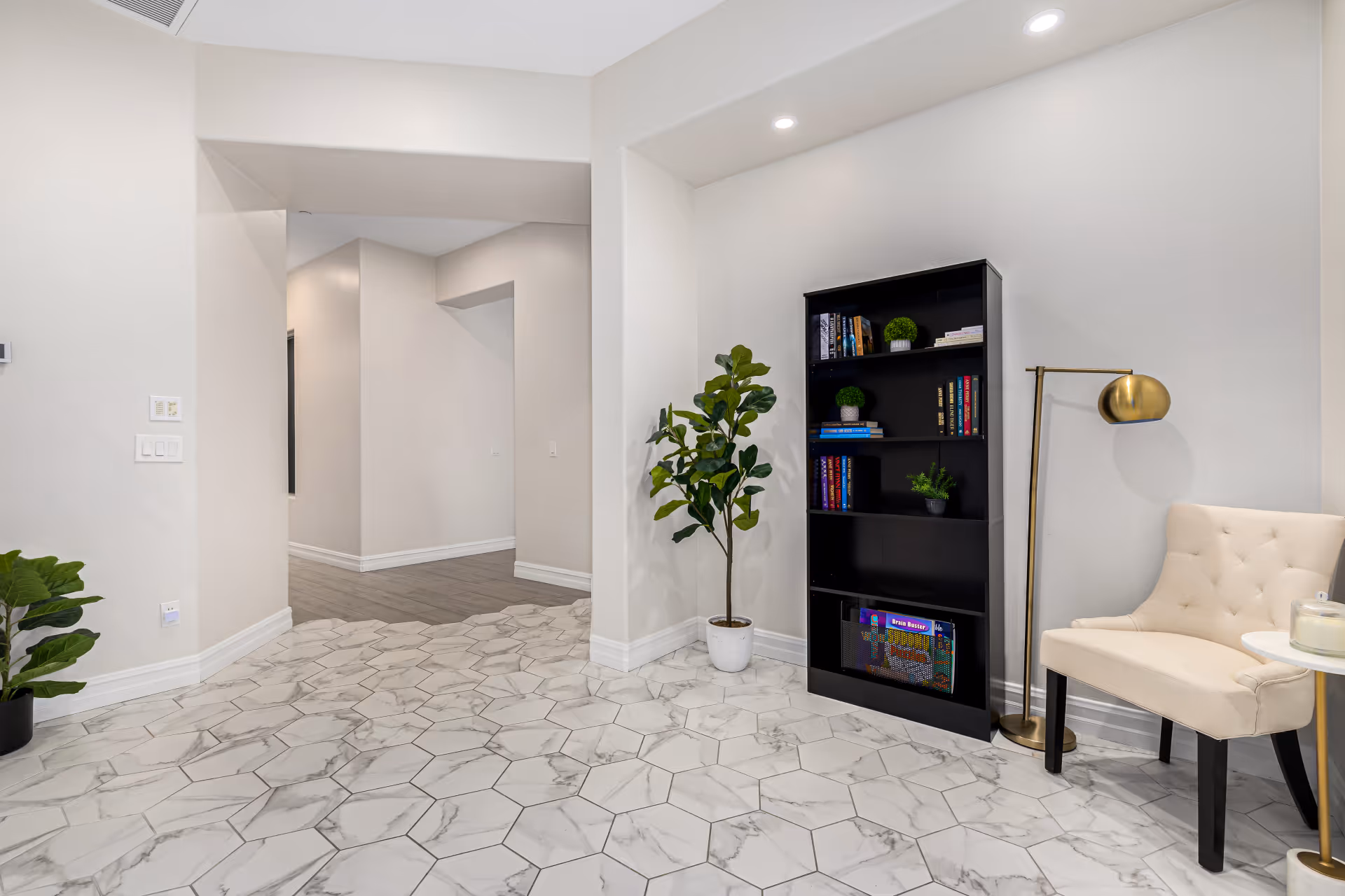 A modern interior space with hexagonal marble floor tiles, a black bookshelf with books and decorative plants, a beige upholstered chair, a gold floor lamp, and a potted plant. The walls are painted white and there is a hallway leading to another room with wooden flooring.