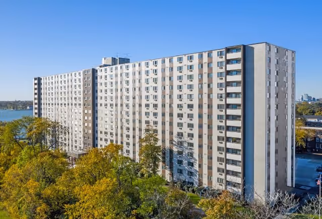 Exterior view of a long multi-story residential building by a river with trees in the foreground under a clear blue sky.