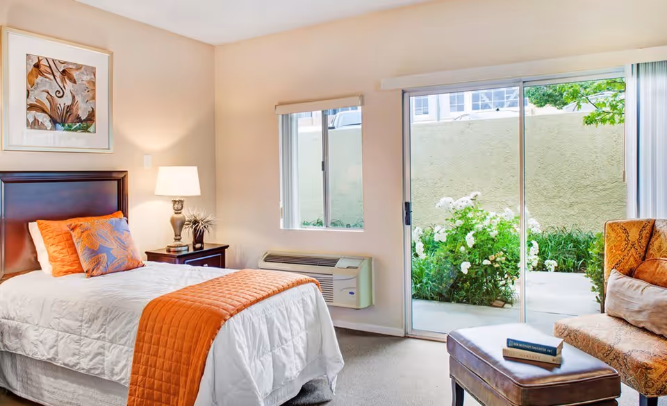 A bright bedroom with a single bed dressed in white bedding and an orange quilt and pillows. Next to the bed is a dark wooden nightstand with a lamp and decorative plant. A framed abstract artwork hangs above the bed. The room has a window and a sliding glass door that opens to a small outdoor garden area with white flowers. There is an upholstered chair with a pillow and a leather ottoman with a book on it near the sliding door.