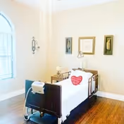 A bright and clean assisted living bedroom with a single hospital-style bed featuring a red heart-shaped pillow. The room has wooden flooring, a large arched window with blinds, a small bedside table with a lamp, and three framed decorative pieces on the wall.