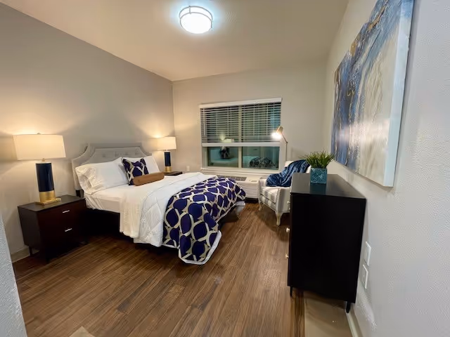 A neatly arranged bedroom with a bed featuring white and blue patterned bedding, two bedside tables with lamps, a window with blinds, a white armchair with a floor lamp beside it, a black dresser with a small plant on top, and a large abstract painting on the wall.