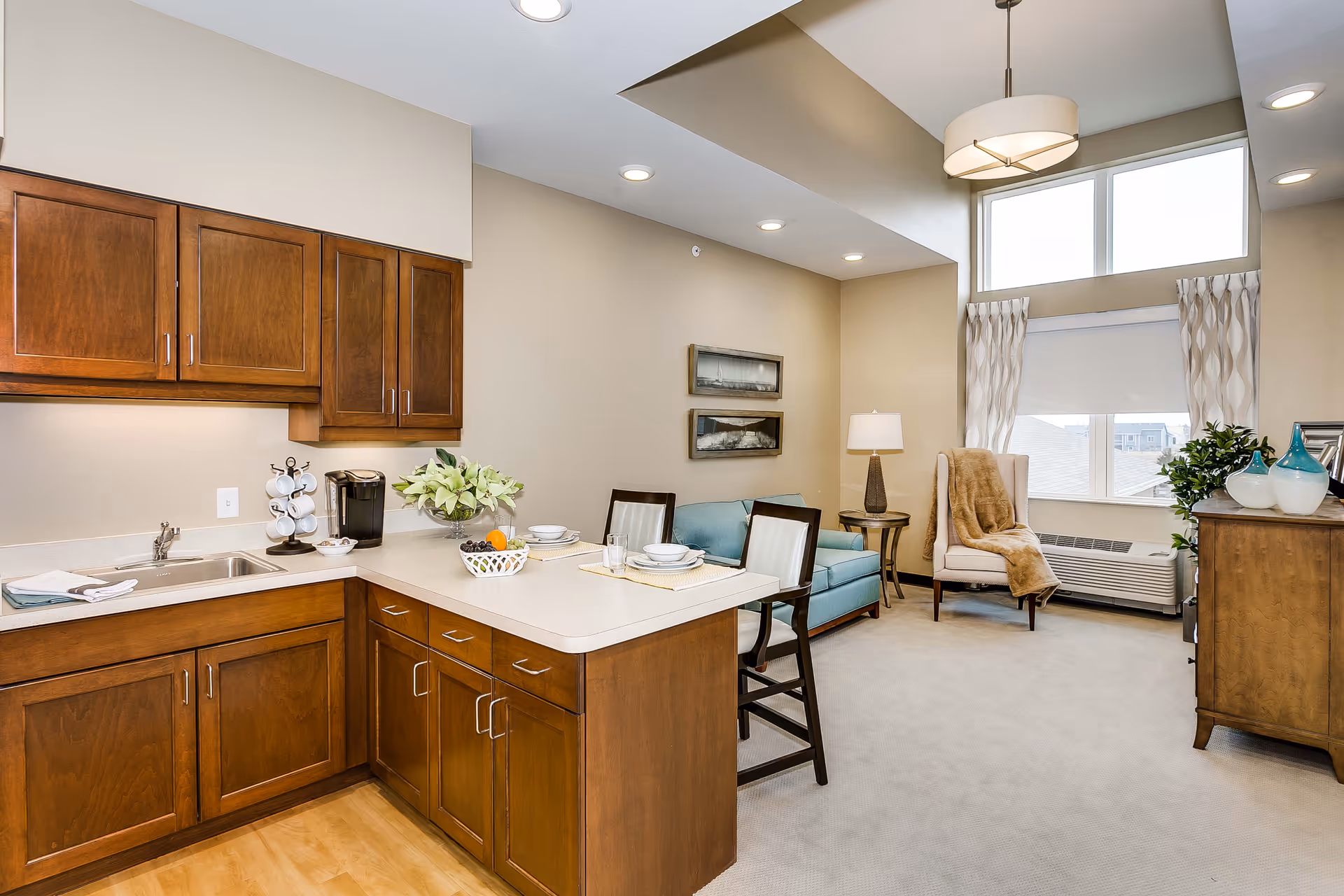 A bright and cozy living space featuring a kitchen area with wooden cabinets and a countertop with two place settings, a coffee maker, and a bowl of fruit. Adjacent to the kitchen is a small living room with a light blue loveseat, a beige armchair draped with a brown throw, a side table with a lamp, and a large window with patterned curtains letting in natural light.
