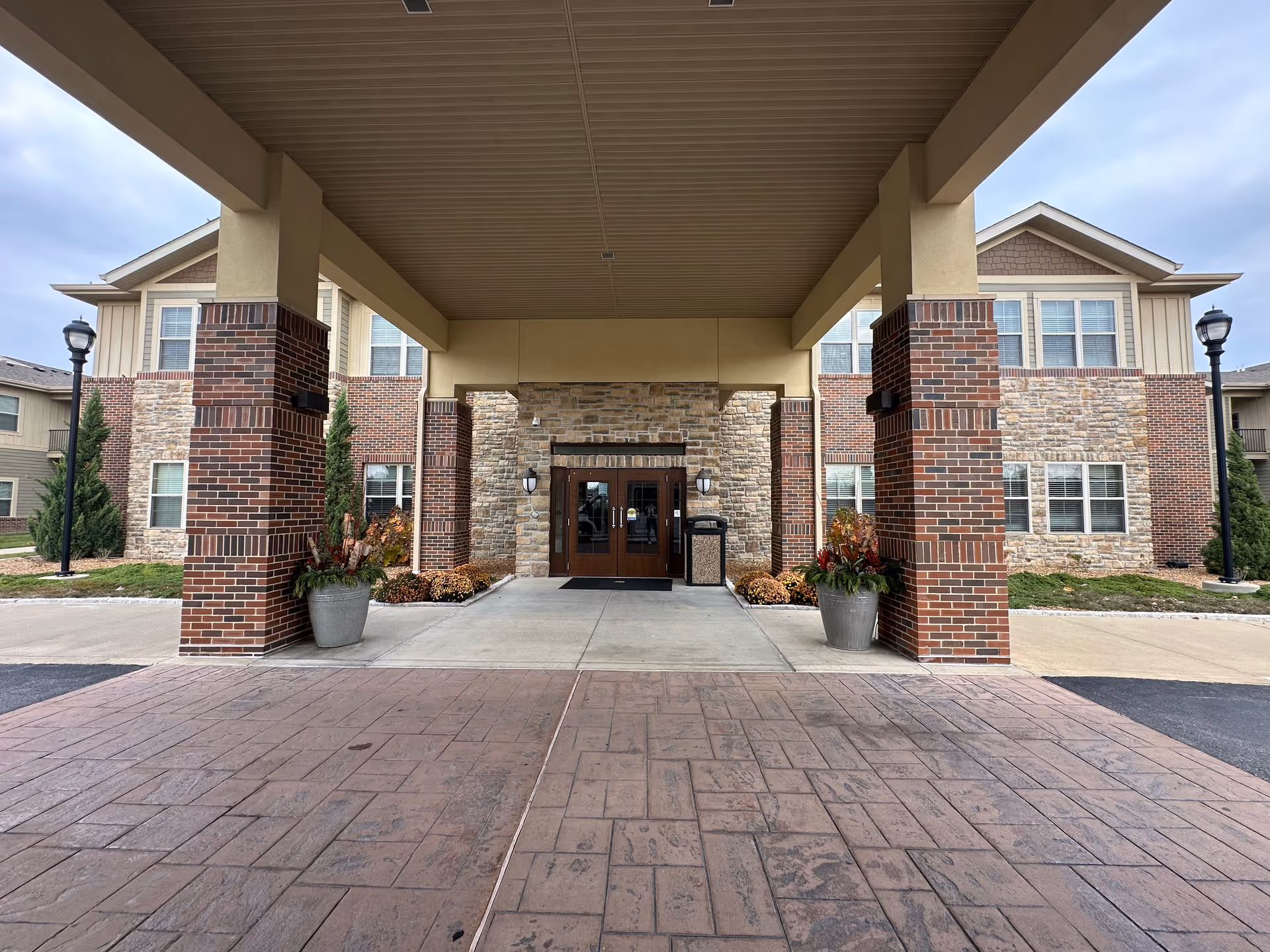 Entrance of Boulevard Senior Living Of St Charles showing a covered driveway with brick and stone pillars, potted plants, and double glass doors leading into the building.