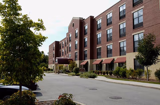 Exterior view of a multi-story brick building with multiple windows and red awnings above the ground floor windows. There is a covered entrance with a maroon canopy and surrounding greenery including trees and shrubs along the sidewalk and parking area.