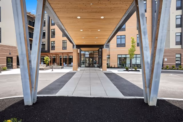 Entrance of The Summit of Blue Ash facility showing a covered walkway with wooden ceiling supported by metal beams, leading to glass doors of a multi-story building with brick and beige exterior walls, surrounded by small trees and landscaping.