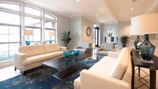 Bright and spacious living room with large windows allowing natural light, featuring two white sofas, a dark coffee table with decorative blue vases, a patterned blue rug, and various plants and decorative items in the background.