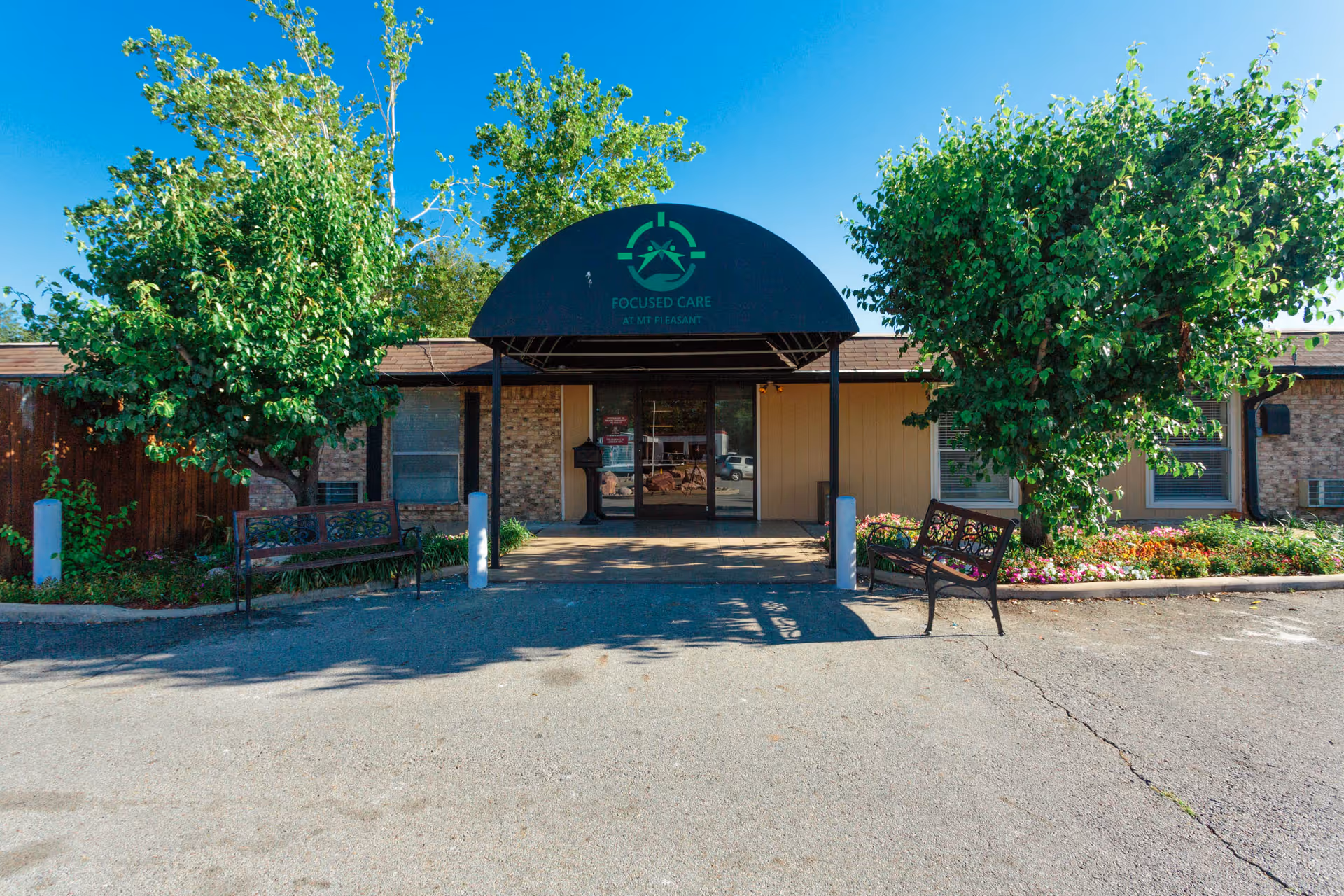 Front entrance of Focused Care at Mount Pleasant facility with a black awning displaying the facility's logo and name. There are two benches on either side of the entrance, flanked by green trees and flower beds. The building exterior is made of brick and wood paneling under a clear blue sky.