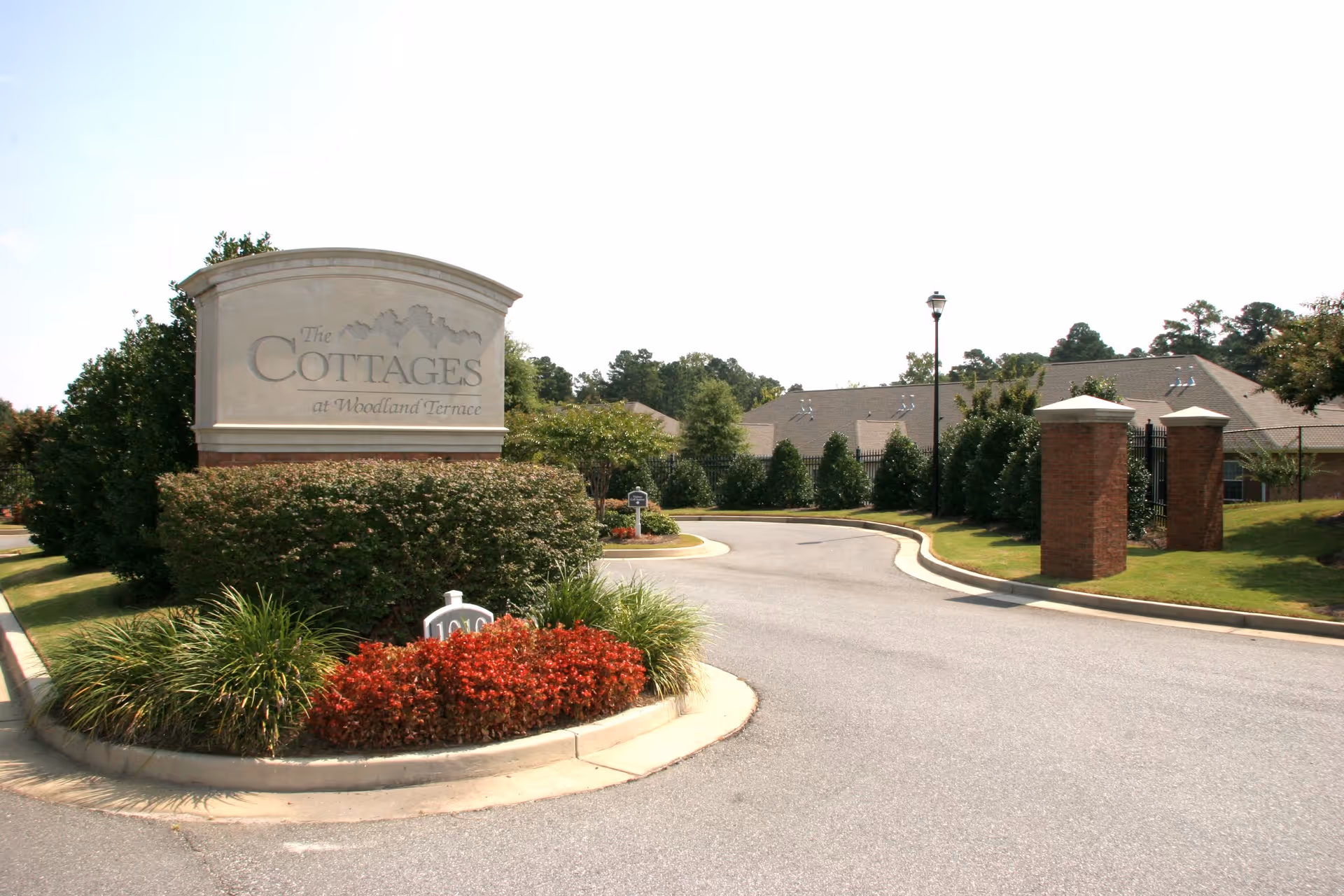 Entrance to The Cottages at Woodland Terrace senior living facility with a large stone sign surrounded by bushes and flowers, a curved driveway, and buildings and trees in the background under a clear sky.
