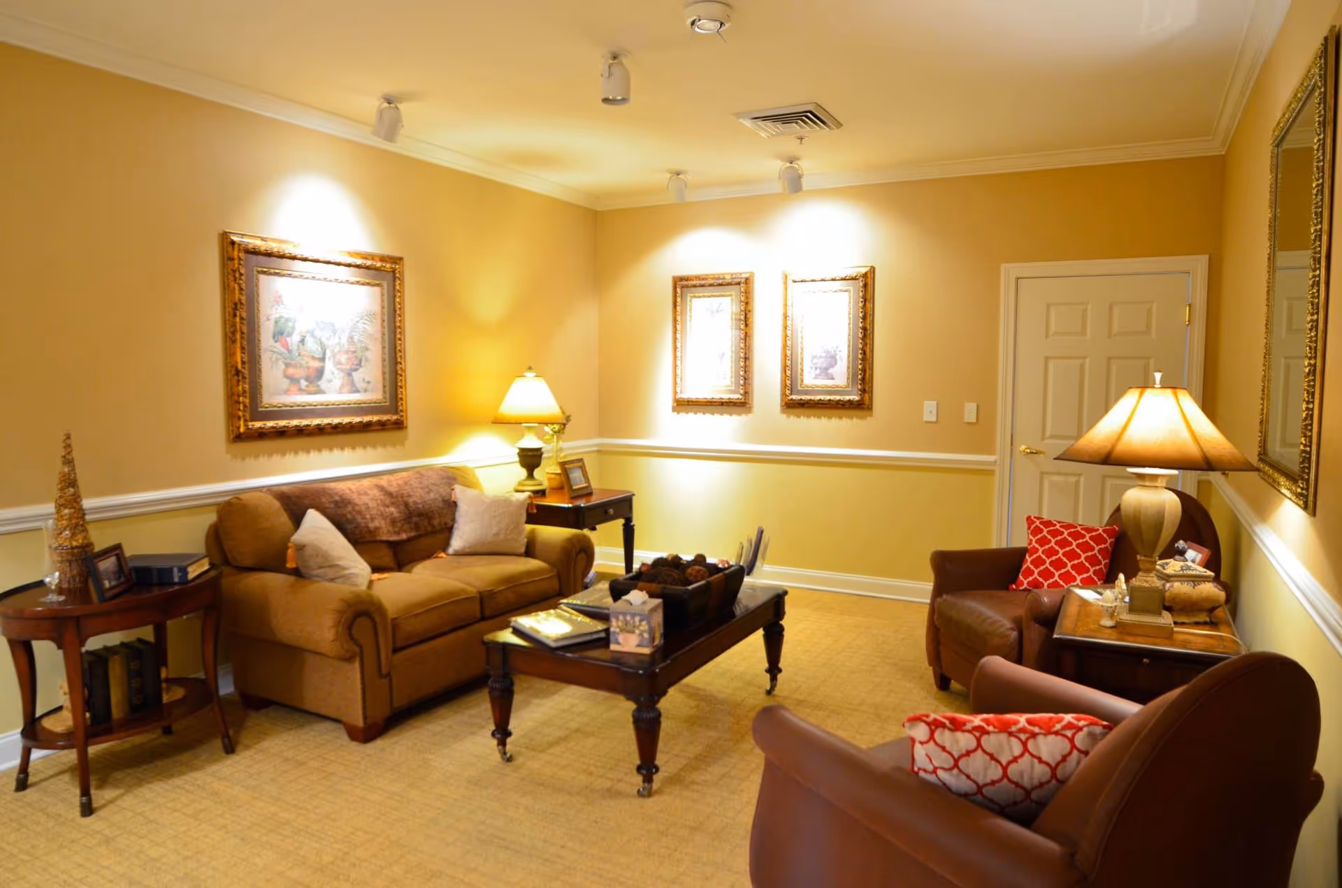 A cozy living room with beige walls and carpet, featuring a brown sofa with pillows, two brown armchairs with red patterned pillows, a wooden coffee table with decorative items, two side tables with lamps, framed artwork on the walls, and a closed white door.