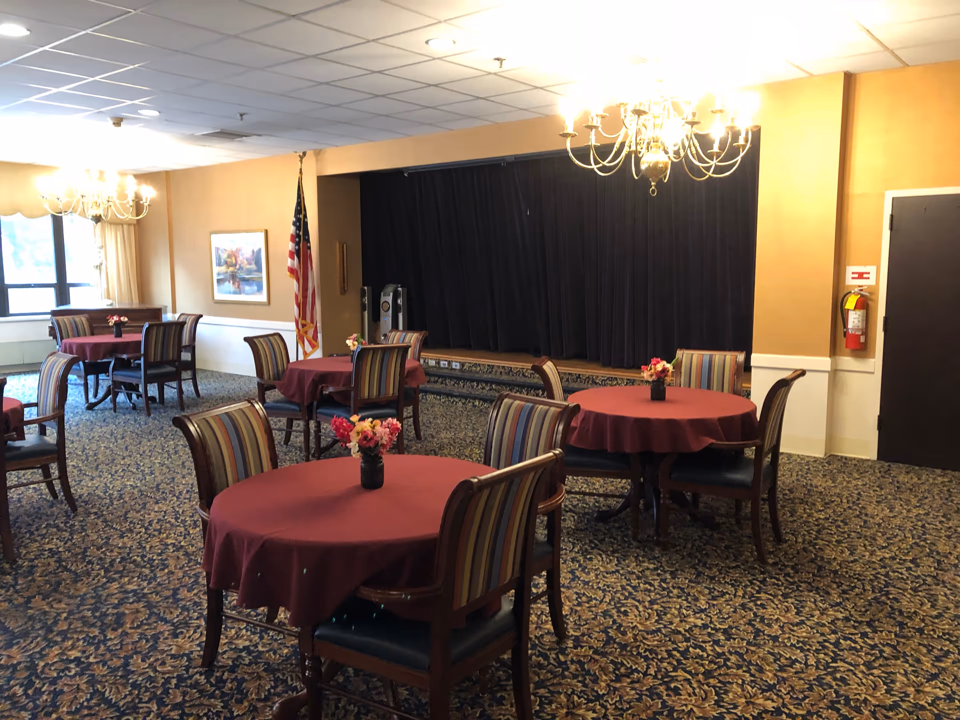 A dining room with round tables covered in burgundy tablecloths, each table having a small flower arrangement. The chairs have striped upholstery. There are chandeliers hanging from the ceiling, a stage with a black curtain in the background, an American flag, and a fire extinguisher mounted on the wall.