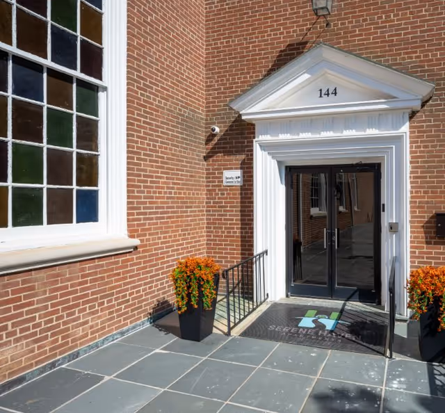 Entrance to a brick building with the number 144 above the white-trimmed doorway. There are two black planters with orange flowers on either side of the entrance, a security camera mounted on the wall, and a mat on the ground with the Serene Haven Apartments logo.