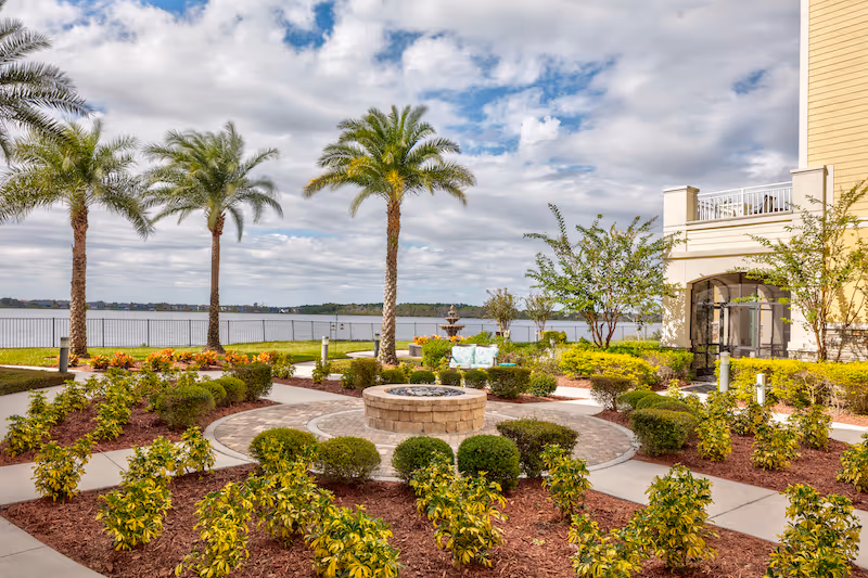 Outdoor garden area with a circular stone fire pit surrounded by neatly trimmed bushes and plants, palm trees, a paved walkway, and a view of a large body of water under a partly cloudy sky. Part of a yellow building with a balcony is visible on the right side.