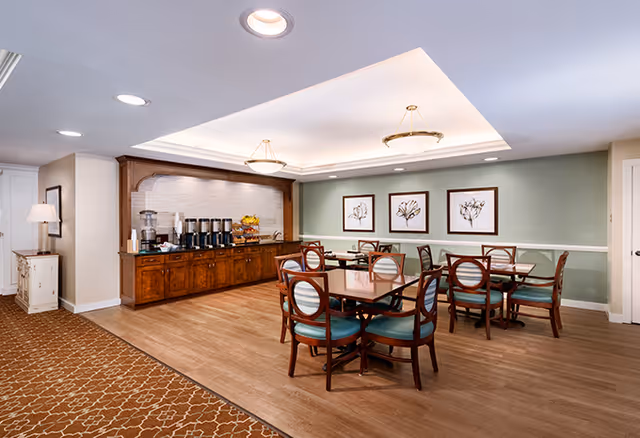 A dining area in a senior living facility with wooden tables and chairs featuring teal cushions. On the left side, there is a wooden buffet counter with coffee dispensers, cups, and a fruit basket. The walls are painted light green with white trim and decorated with three framed botanical prints. The ceiling has recessed lighting and two hanging light fixtures. The floor is a combination of wood and patterned carpet.