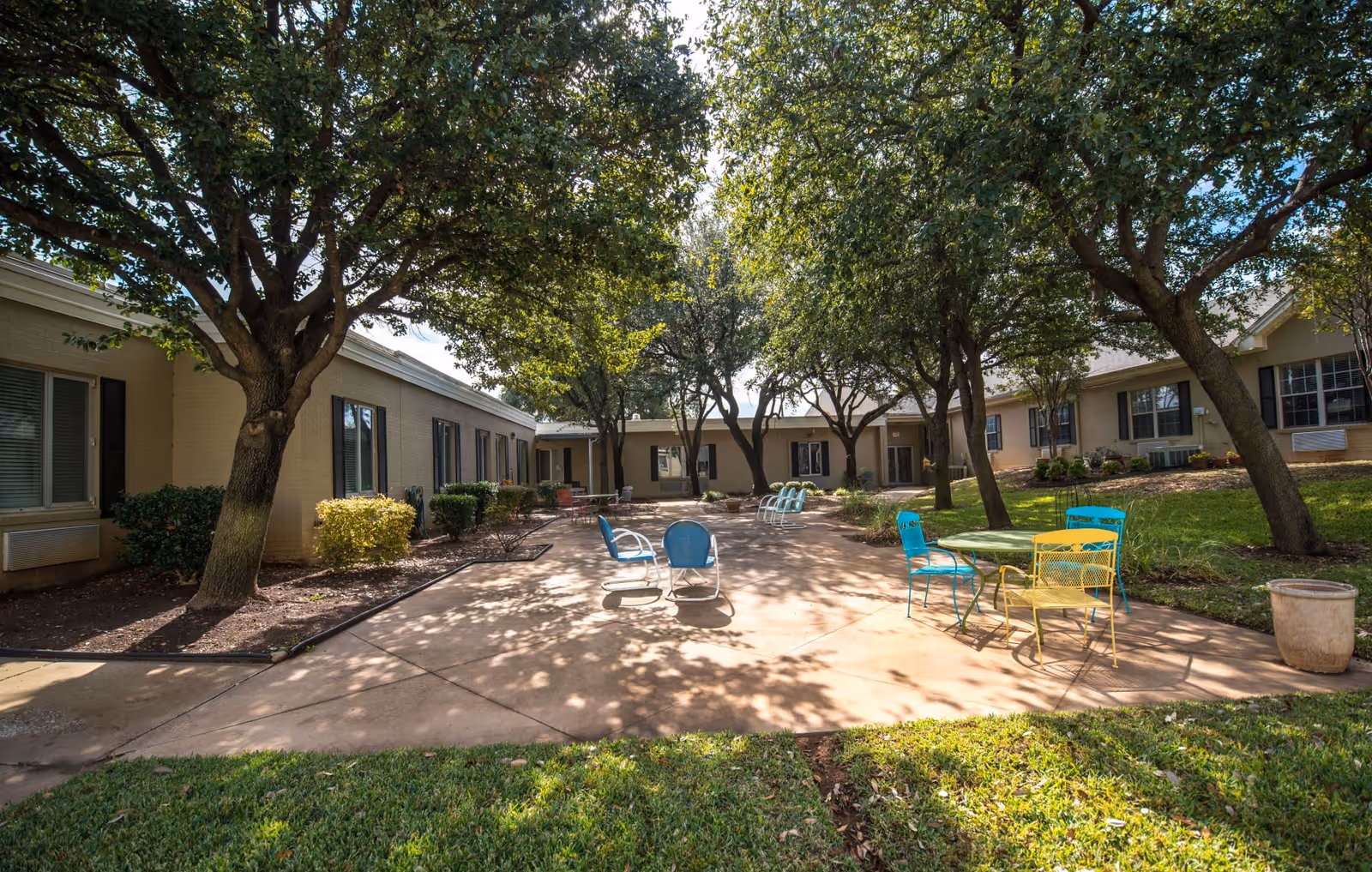 Outdoor courtyard area at Ashwood Court featuring a paved walkway surrounded by single-story buildings with windows. Several trees provide shade over the area, and there are colorful metal chairs and tables arranged on the pavement. The grass and landscaping are well-maintained under a partly cloudy sky.