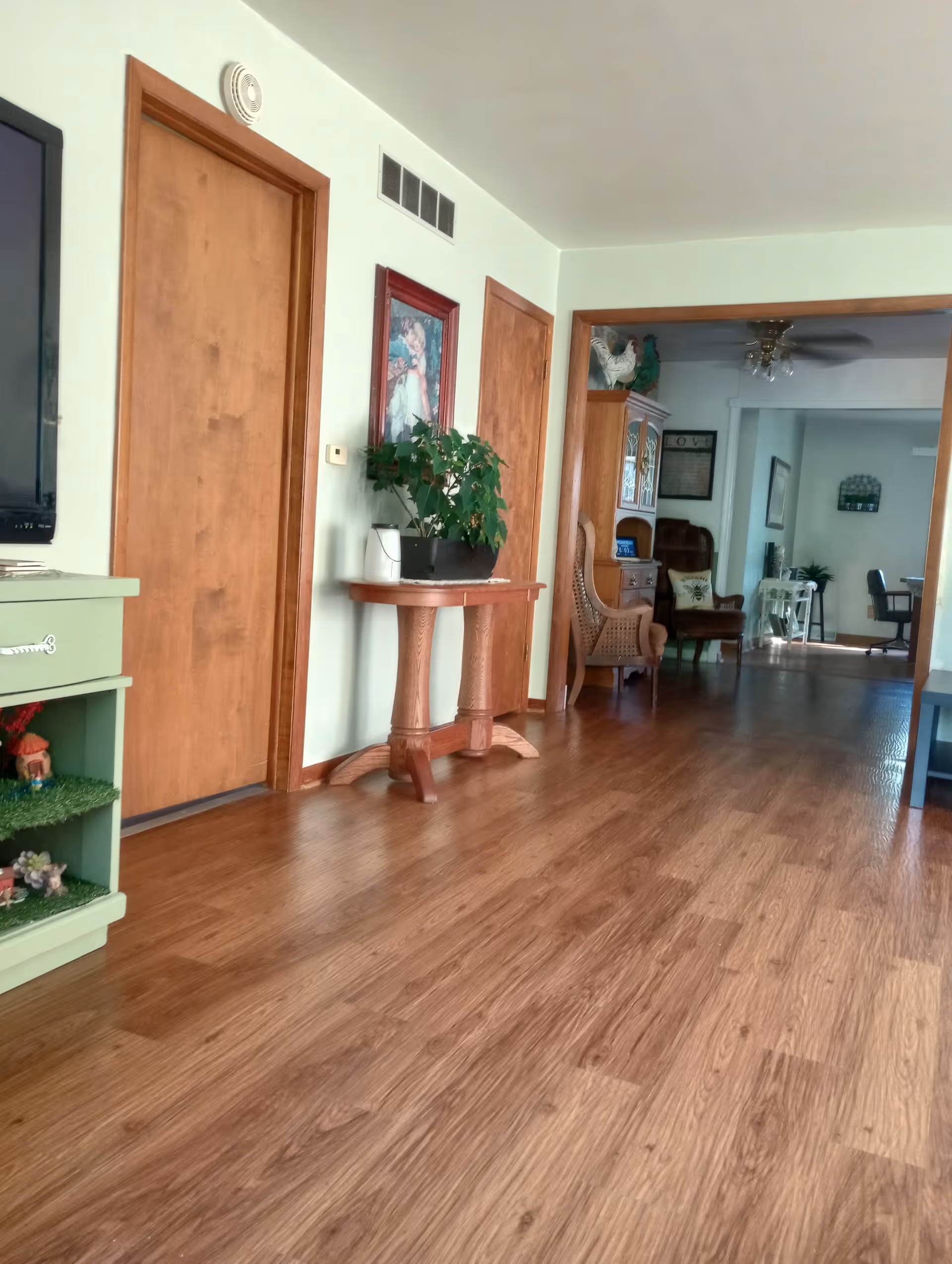 Wood‑floored interior hallway and living area with wooden doors, a small table holding a potted plant, and seating visible in the adjoining room.