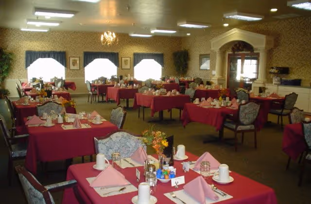 A dining room with many tables covered in burgundy tablecloths, set with plates, cups, folded napkins, and floral centerpieces.