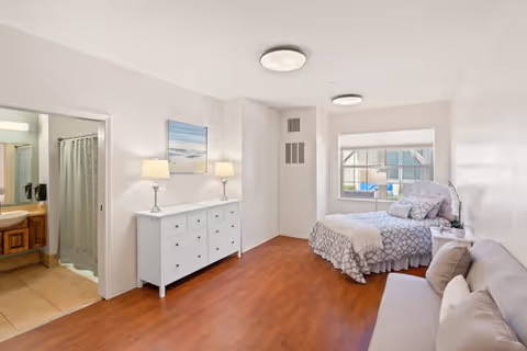 A bright and clean senior living bedroom featuring a single bed with patterned bedding near a window, a white dresser with two lamps and a beach-themed painting above it, a beige sofa with pillows, and an open door leading to a bathroom with a shower curtain.