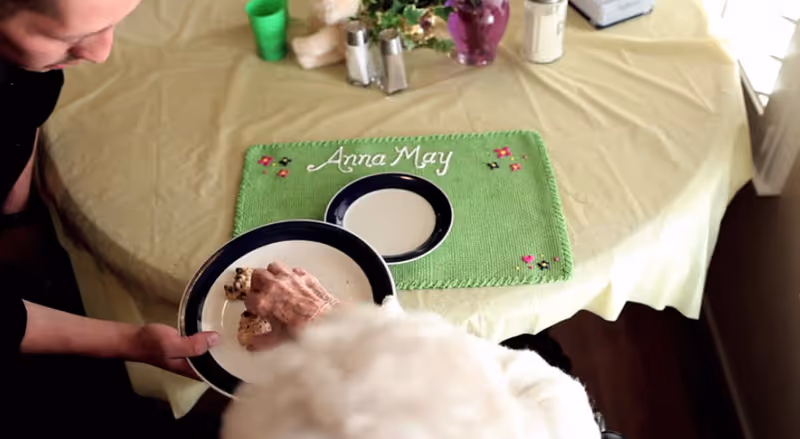 An elderly person reaching for food on a plate held by another person at a table with a green placemat embroidered with the name 'Anna May'. The table is covered with a light yellow tablecloth and has a small flower arrangement and salt and pepper shakers.
