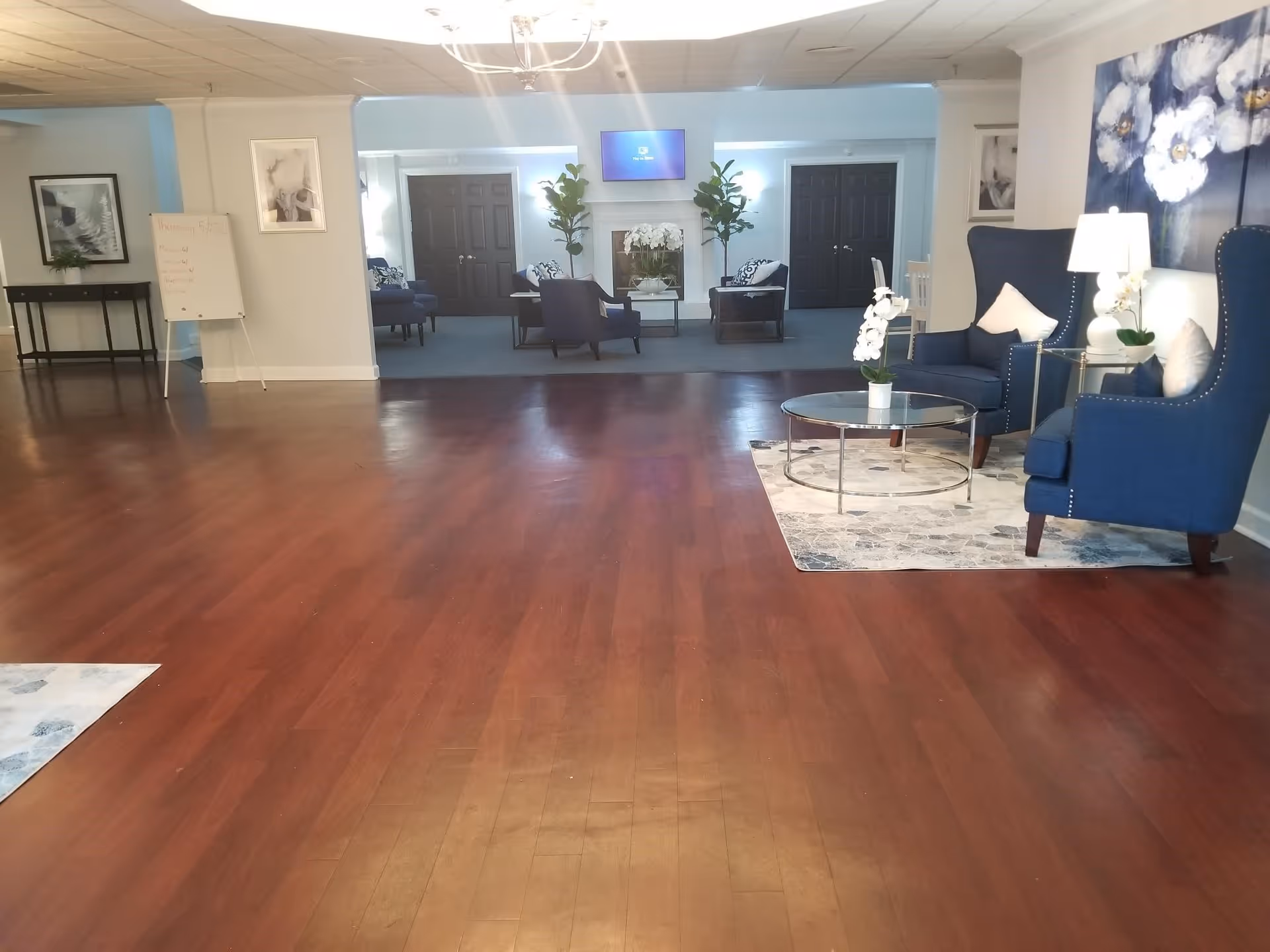 A spacious senior living facility common area with polished wooden floors and a seating area on the right featuring two blue armchairs, a glass coffee table on a patterned rug, and a lamp. In the background, there is another seating area with blue chairs, a fireplace, two tall plants, and a wall-mounted TV. The walls are light-colored with framed artwork and a whiteboard on the left side.