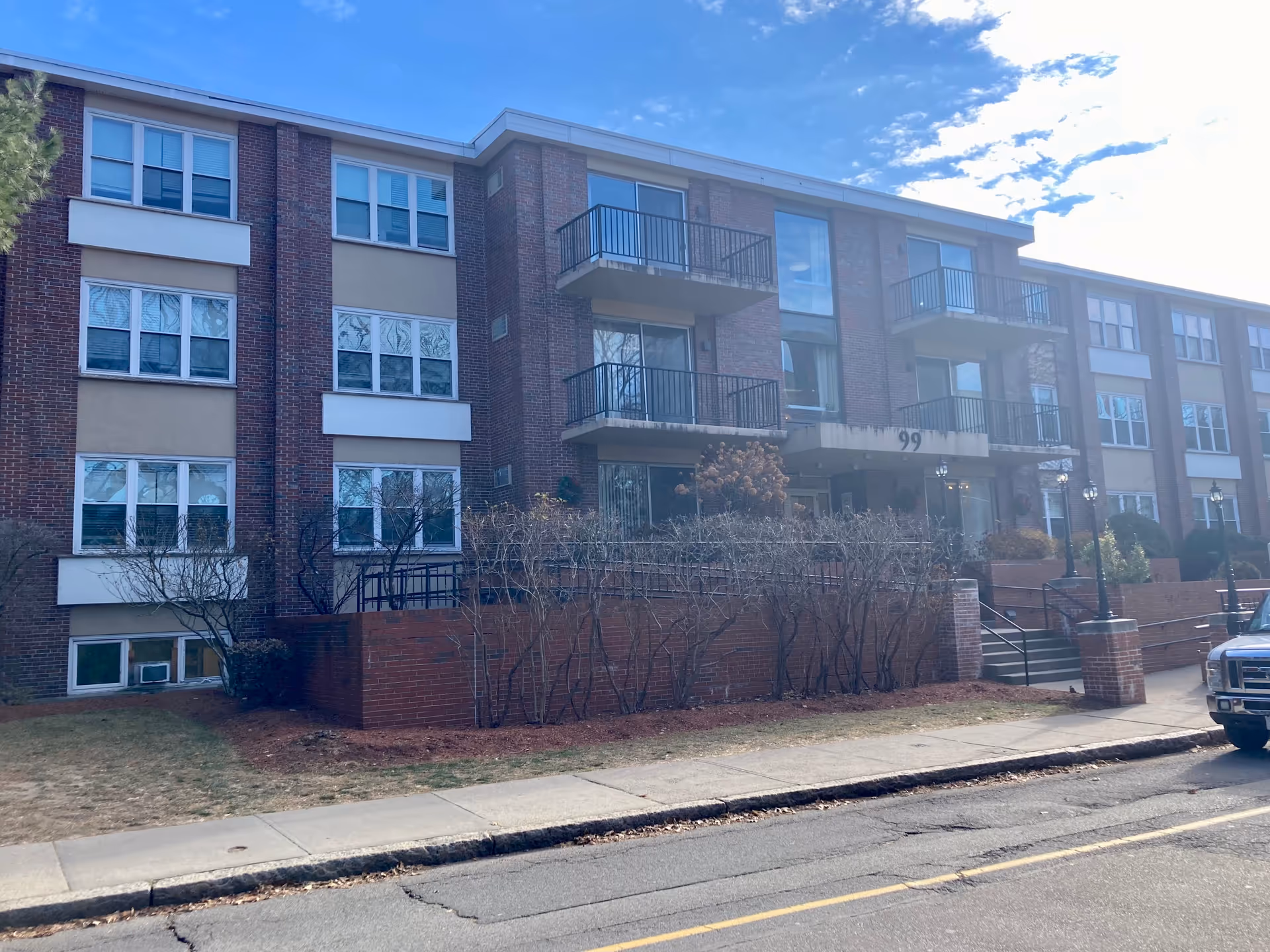 Exterior view of a multi-story brick and beige senior living facility building with balconies, windows, and a main entrance with stairs and railings. There are leafless bushes along the front and a parked vehicle on the street in front of the building under a partly cloudy sky.