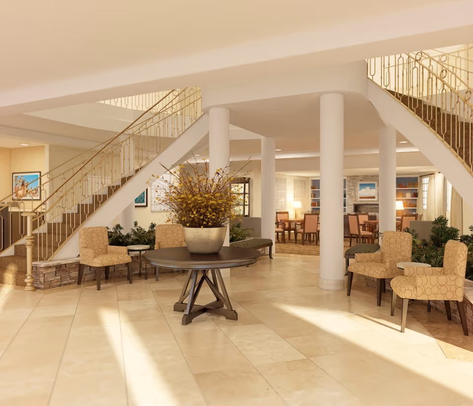 A spacious and well-lit senior living facility common area with beige tiled floors, a round table with a large flower arrangement in the center, several patterned armchairs, and a staircase with a decorative gold railing. In the background, there are tables and chairs near a stone fireplace and bookshelves, creating a cozy lounge atmosphere.