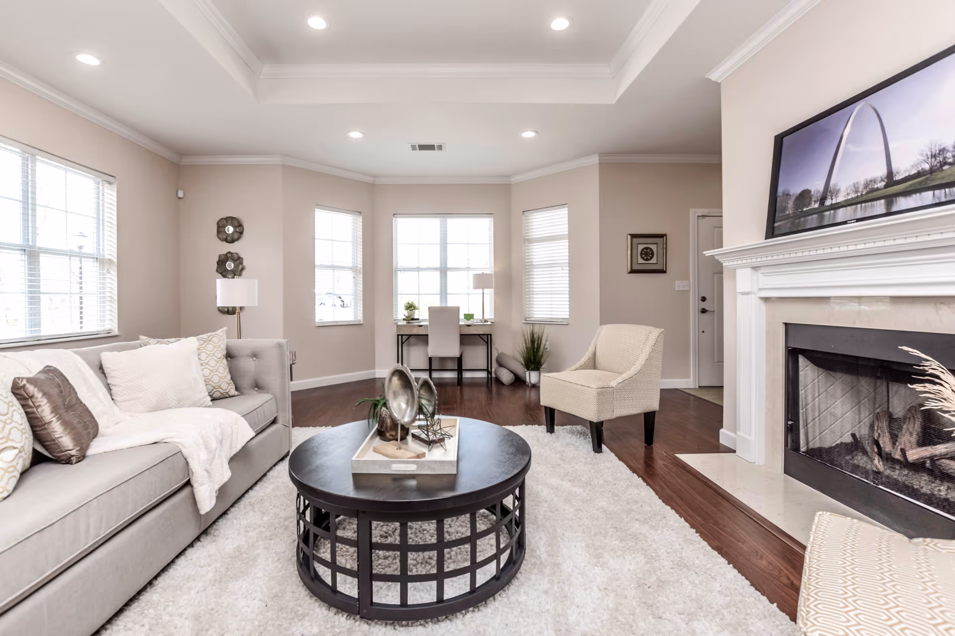 A bright living room with a gray sofa adorned with pillows and a throw blanket, a round black coffee table with decorative items, a beige armchair, a white fluffy rug, a fireplace with a framed picture above it, and a small desk with a chair near three windows.
