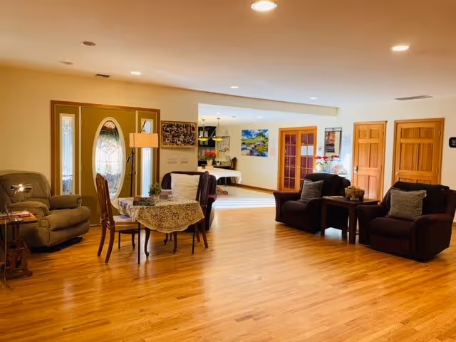 A spacious living room area with wooden flooring, featuring a round table with a lace tablecloth and four chairs, two dark armchairs with cushions, a recliner chair, and a floor lamp near a decorative front door with glass panels. The room is well-lit with recessed ceiling lights and has a view into an adjacent dining area with a table and chairs.