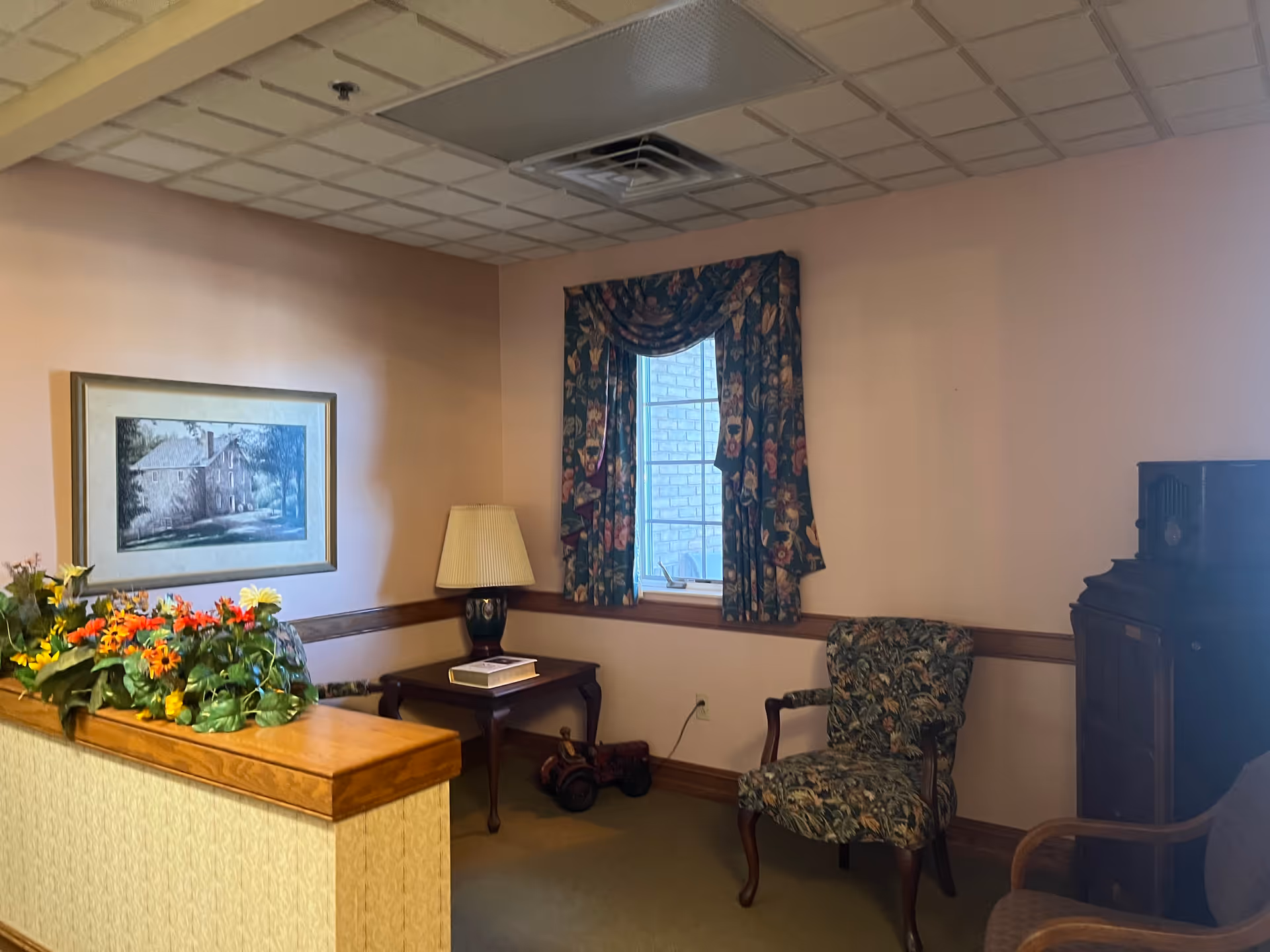 A cozy corner of an interior room with peach-colored walls, a window with floral curtains, a floral upholstered armchair, a wooden side table with a lamp and a book, a framed painting on the wall, and a decorative arrangement of artificial flowers on a half wall.
