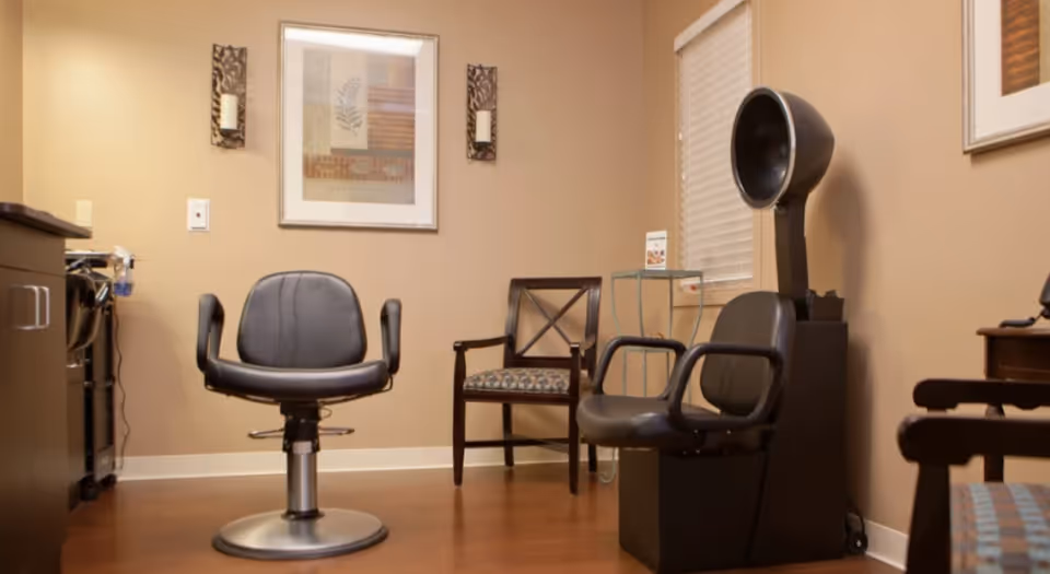Interior view of a salon area with a black salon chair in the center, a wooden chair with patterned upholstery, a hair dryer chair with a hood dryer, and framed artwork on beige walls.