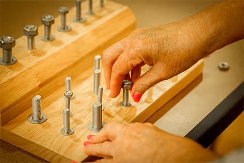 Close-up of an elderly person's hands engaging with a wooden peg and bolt activity, likely used for hand therapy or rehabilitation exercises.