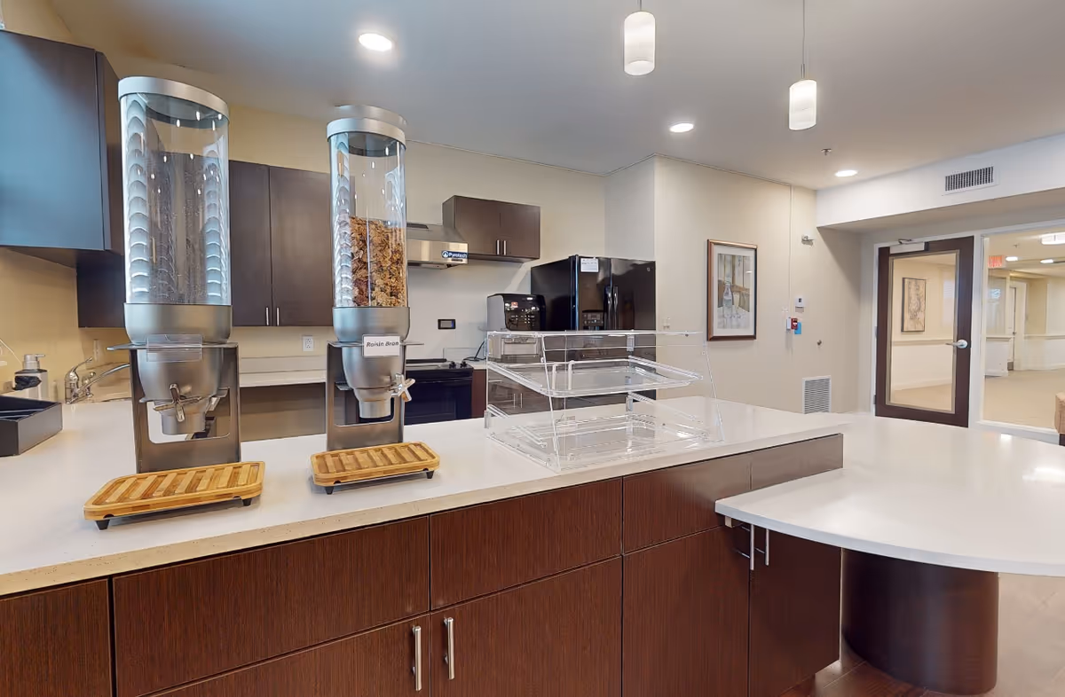 Interior view of a senior living facility kitchen area with dark wood cabinets, a white countertop island, two cereal dispensers labeled Raisin Bran, a clear plastic display case, and kitchen appliances including a refrigerator and stove. The area is well-lit with ceiling lights and pendant lights, and there is a door leading to a hallway in the background.