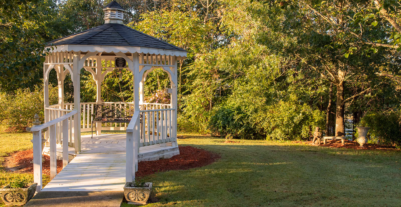 A white wooden gazebo with a bench on a sunlit grassy lawn surrounded by trees and shrubs.