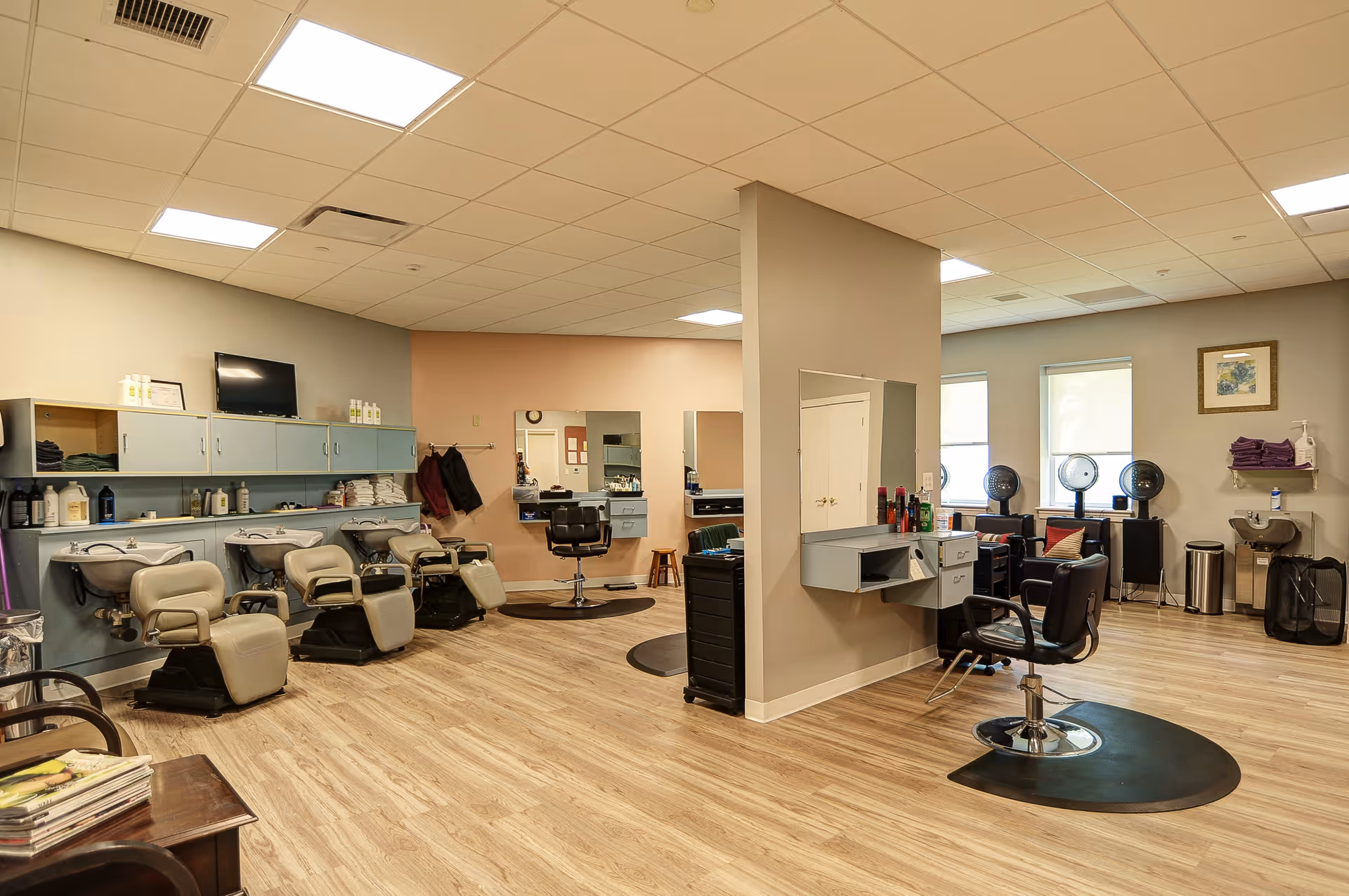 Interior view of a hair salon area in a senior living facility with multiple salon chairs, hair washing stations, mirrors, and hair dryers. The room has wood flooring, beige and light pink walls, and ceiling lights.
