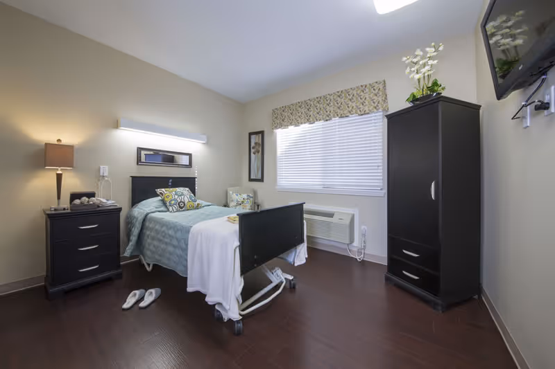 A tidy furnished nursing room with a single hospital-style bed, nightstand, wardrobe, and wall-mounted TV.