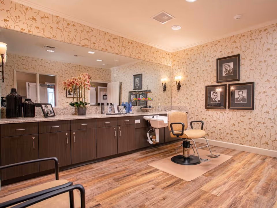A well-lit salon area with a single beige salon chair on a mat in front of a large mirror. The room has floral wallpaper, wooden flooring, and a long countertop with cabinets underneath. On the countertop are decorative items including a vase with flowers, framed photos, and various hair care products. Three framed black-and-white portraits hang on the wall to the right.