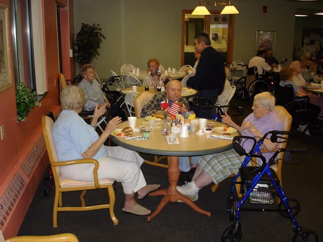 Several elderly residents sitting at round tables in a dining area of an assisted living community, eating and conversing. Some residents use walkers and wheelchairs. The room has warm lighting and a cozy atmosphere.