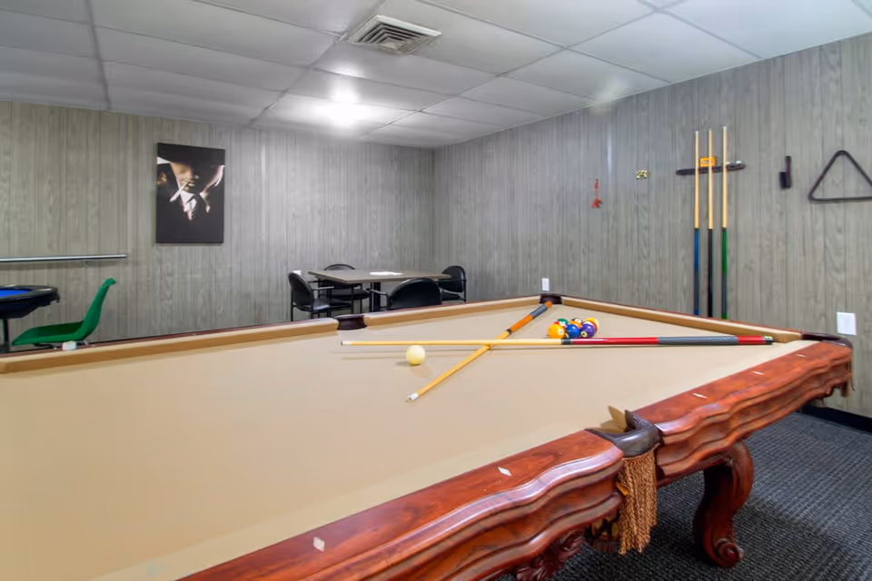 A game room with a beige felt pool table in the foreground, pool cues resting on the table, and a rack of cues mounted on the wood-paneled wall. In the background, there is a small table with four chairs and a framed picture on the wall. The room has a drop ceiling with fluorescent lighting.