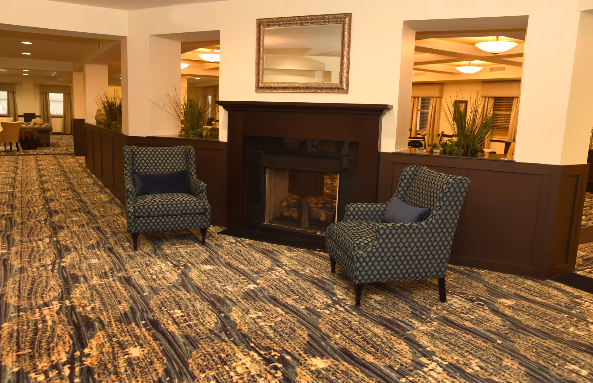 A cozy interior lounge area with two patterned armchairs facing a dark wooden fireplace. Above the fireplace is a large framed mirror. The room features a patterned carpet and warm lighting, with plants placed on a wooden divider behind the chairs.
