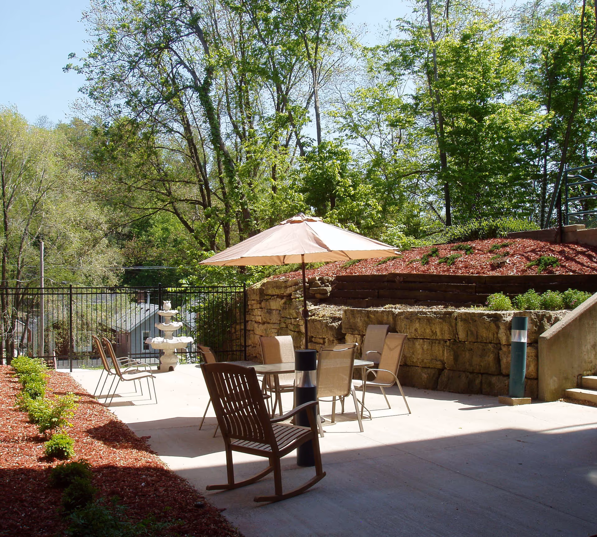 Outdoor patio area with a table and four chairs under a beige umbrella, a wooden rocking chair, and a multi-tiered white fountain near a black metal fence. The patio is surrounded by landscaped areas with mulch, small bushes, and large stone retaining walls, with trees and greenery in the background under a clear blue sky.