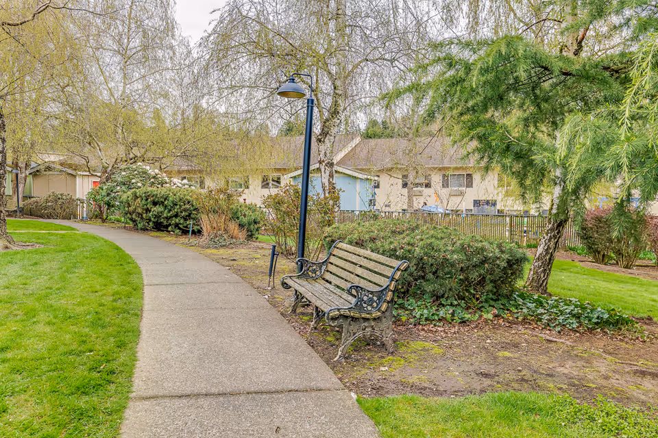A paved walking path curves through a landscaped garden area with green grass, bushes, and trees. A wooden bench with ornate metal armrests and legs is positioned next to a black lamppost along the path. In the background, there are single-story buildings with beige and blue walls and windows.