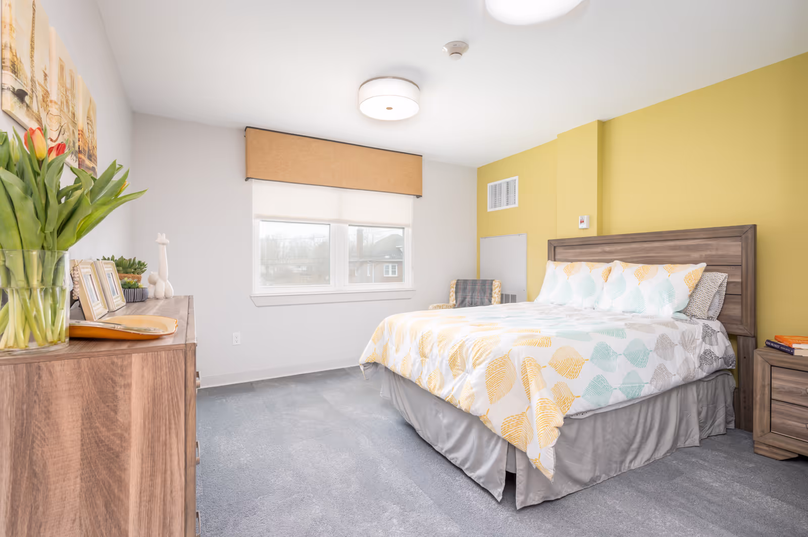 A bright and clean bedroom with a large bed featuring a white comforter with yellow, teal, and gray leaf patterns. The room has a yellow accent wall behind the bed, a wooden headboard, a wooden nightstand with books, and a wooden dresser with framed photos and a vase of tulips. A window with a beige valance lets in natural light, and a cozy chair is placed in the corner.