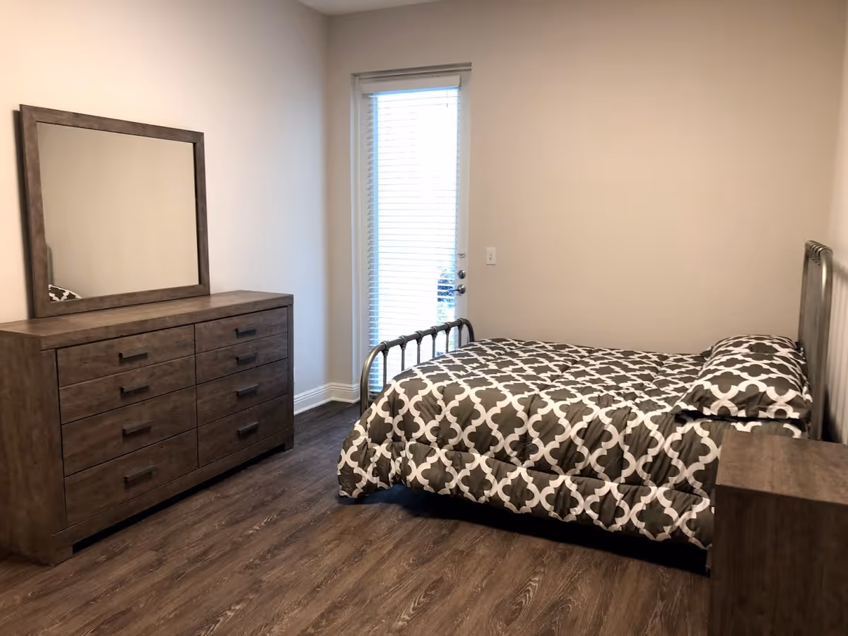 A bedroom with a wooden dresser and mirror on the left, a bed with a patterned black and white comforter and pillow on the right, and a door with blinds in the center background. The room has wood flooring and plain light-colored walls.